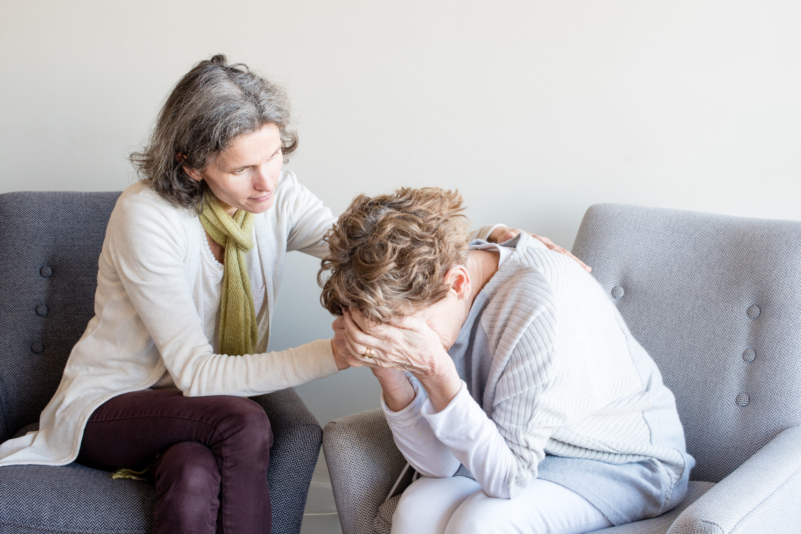 Middle aged daugher in green scarf comforting older woman in cream top with hands over face