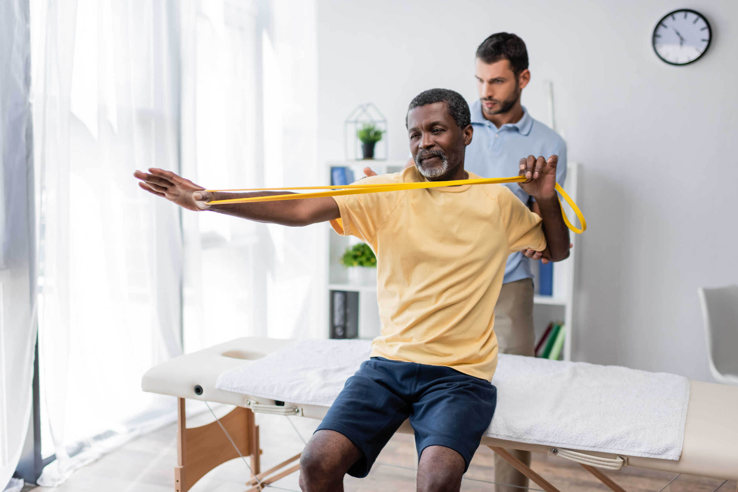 middle aged african american man sitting on massage table and training with elastics near and physical therapy